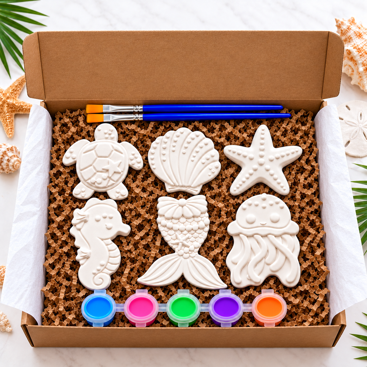 An open cardboard box filled with sea-themed plaster paint items, including white seashell, starfish, seahorse, mermaid tail, and jellyfish and turtle plasters, five paint pots, and two paintbrushes on a light marble surface with starfish and palm leaves around the edges.