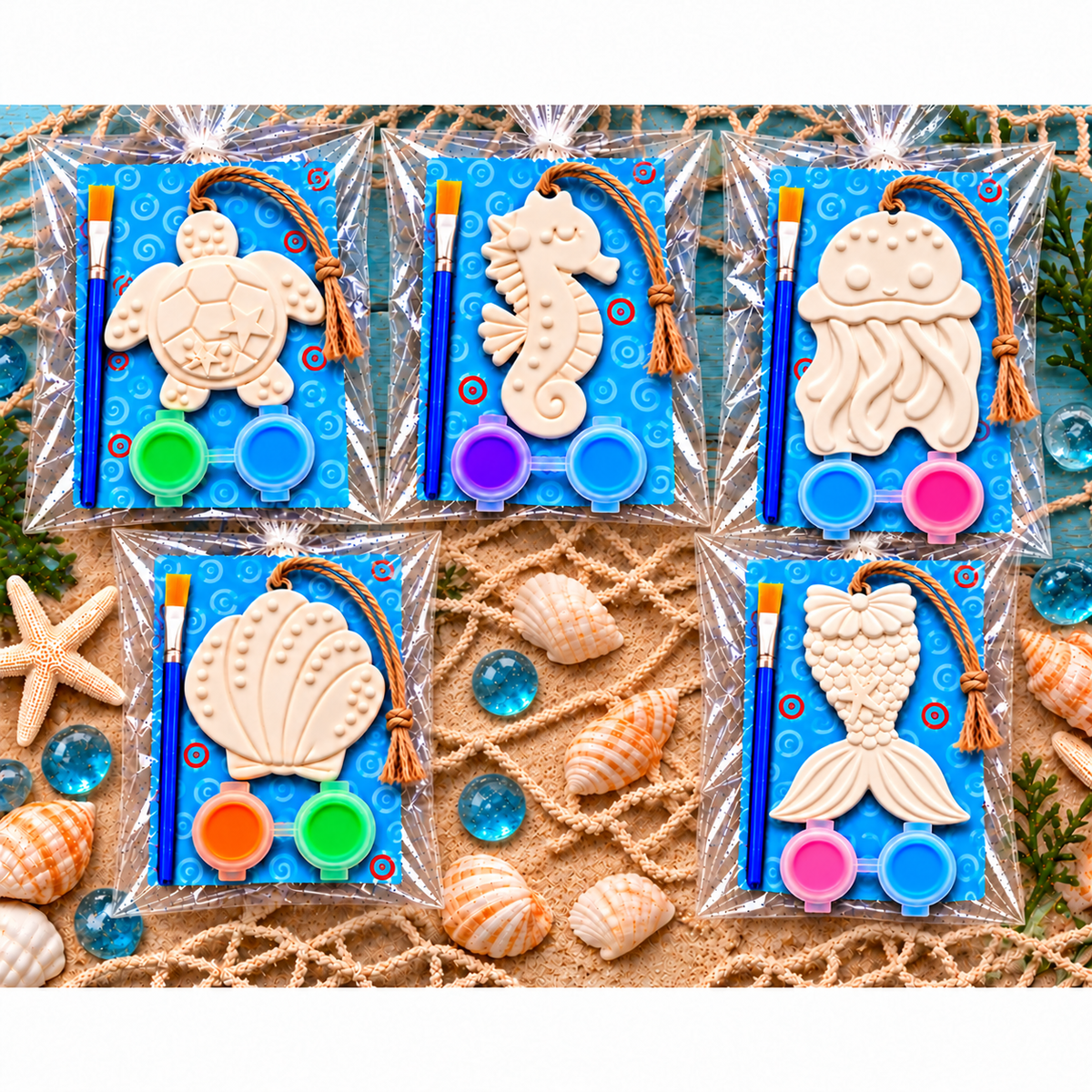 Five packaged sea-themed plaster painting kits arranged on a sandy surface with seashells and decorative props. Each kit includes a plaster sea creature design (turtle, seahorse, jellyfish, clam, mermaid tail) on a blue backing card, a paintbrush, and two small paint pots.