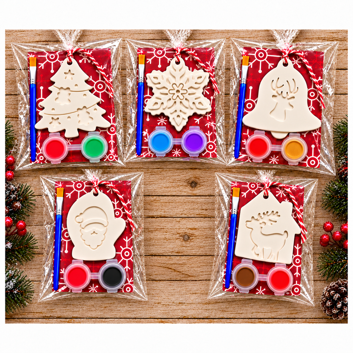 Five Christmas ornament plaster paint kits in clear plastic bags arranged on a wooden surface. Each kit contains an unpainted plaster ornament shape, a red patterned backing card, a blue-handled paintbrush, and small round paint pots.
