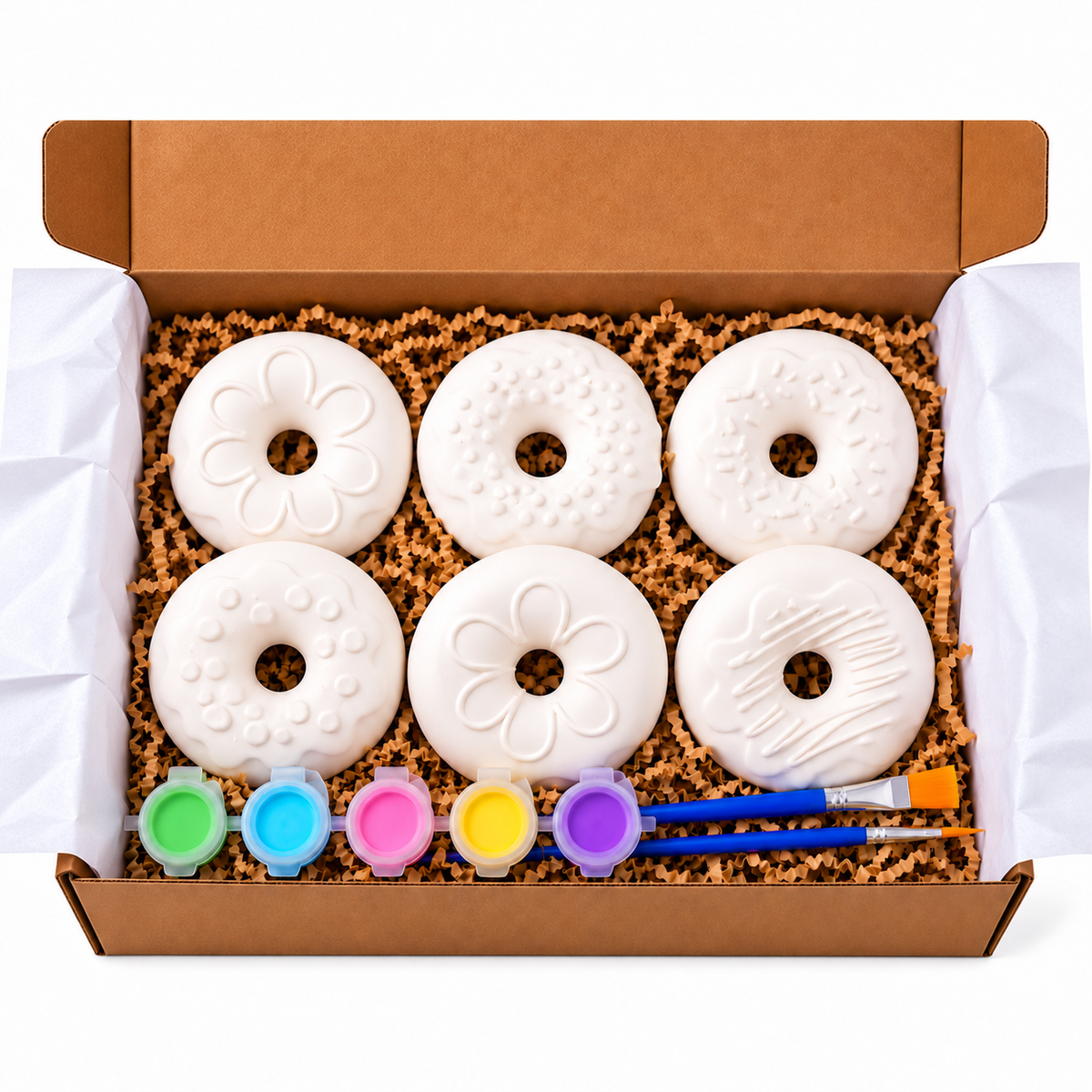 A boxed DIY plaster paint kit featuring six white painted donut-shaped pieces with different embossed and painted patterns, set in brown paper shred inside an open cardboard box. A row of small paint pots in green, blue, pink, yellow, and purple with two blue-handled paintbrushes is included at the bottom.