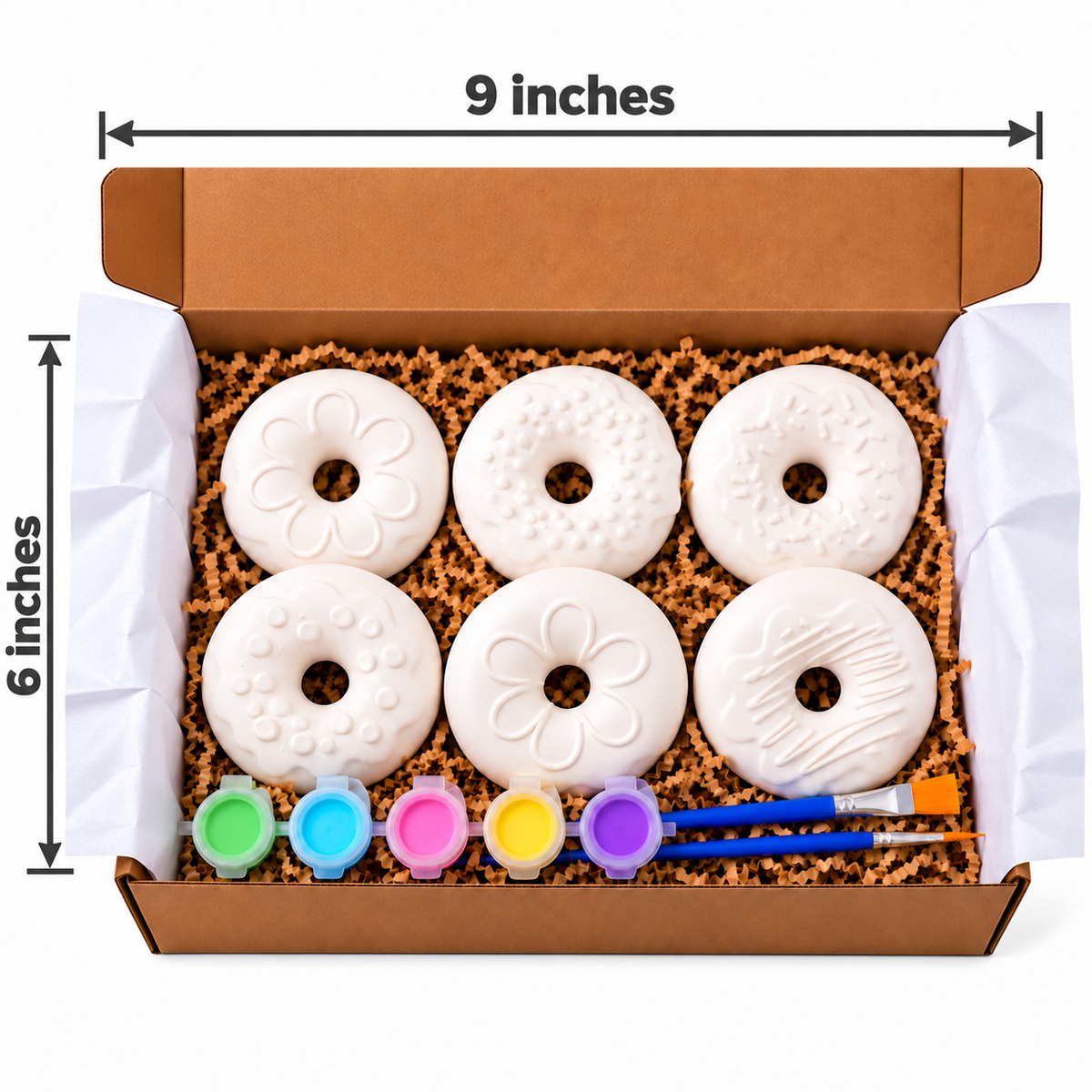 Open cardboard gift box containing six white decorated donut-shaped items in a plaster paint kit, with five small paint pots, two paintbrushes, and dimension labels showing 9 inches by 6 inches on a white background.