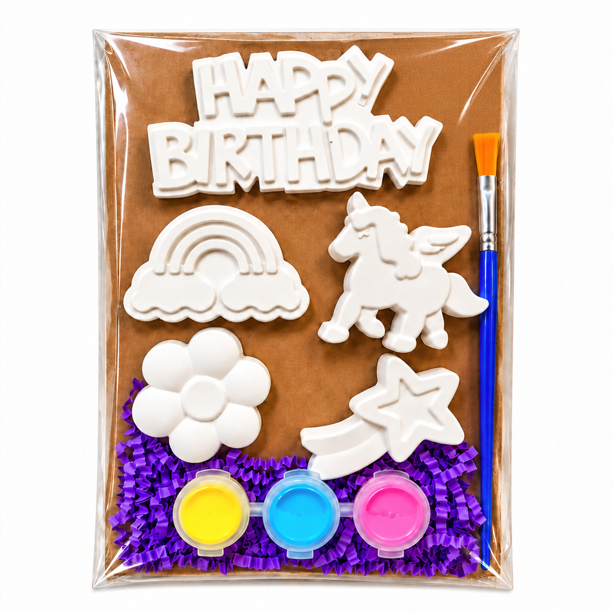 A children's birthday decoration plaster paint kit in clear plastic packaging, featuring white unpainted clay or sugar paste shapes including a unicorn, rainbow, flower, shooting star, and 'HAPPY BIRTHDAY' sign, along with three small paint pots in yellow, blue, and pink and a blue paintbrush on a brown backing card.