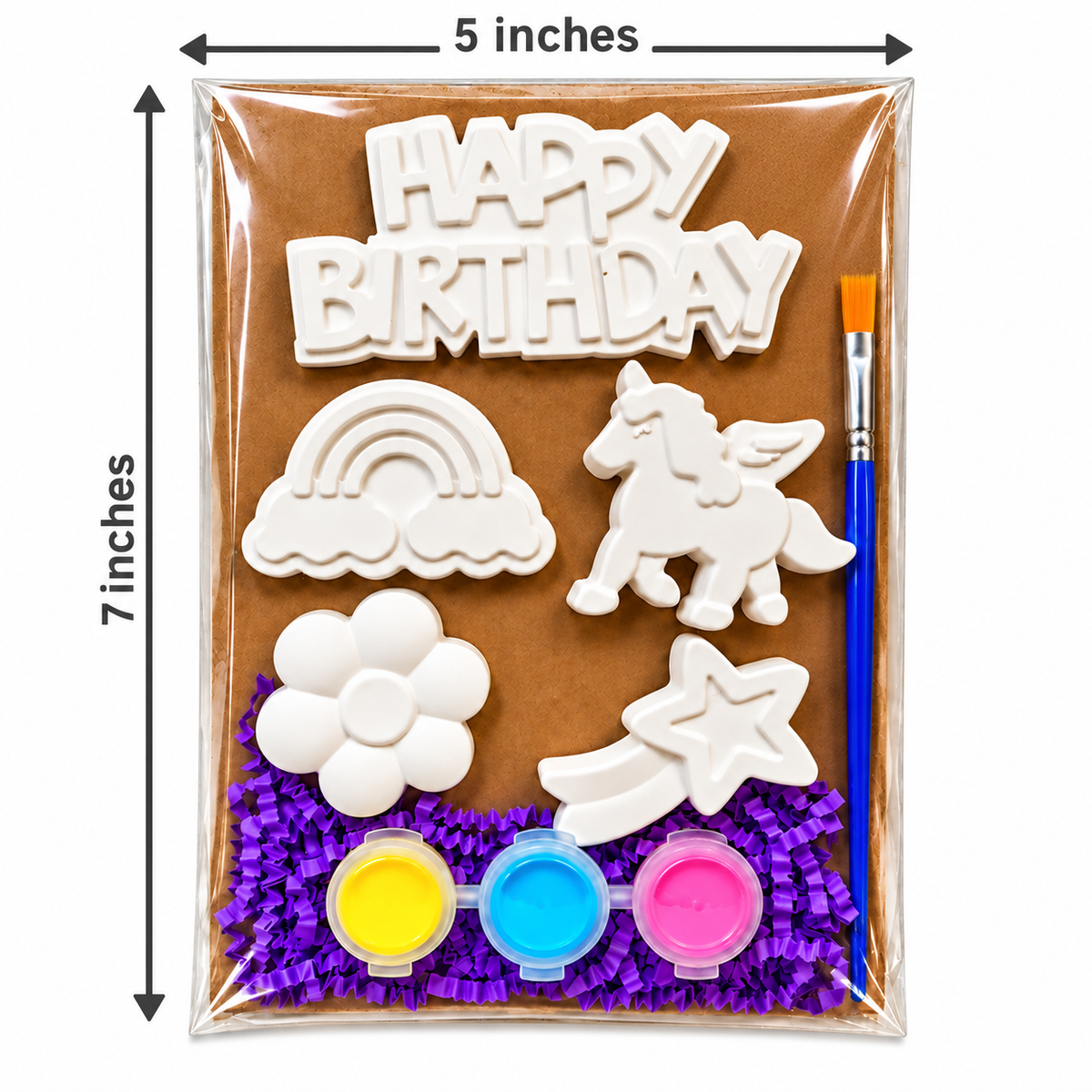 A birthday decoration plaster paint kit in a clear plastic bag with measurement arrows showing 5 inches wide by 7 inches tall. The kit includes white clay or foam cutouts shaped like 'HAPPY BIRTHDAY,' a rainbow, a unicorn, a flower, a shooting star, three small paint pots in yellow, blue, and pink, and a paintbrush, all arranged over purple crinkle paper on a brown backing card.