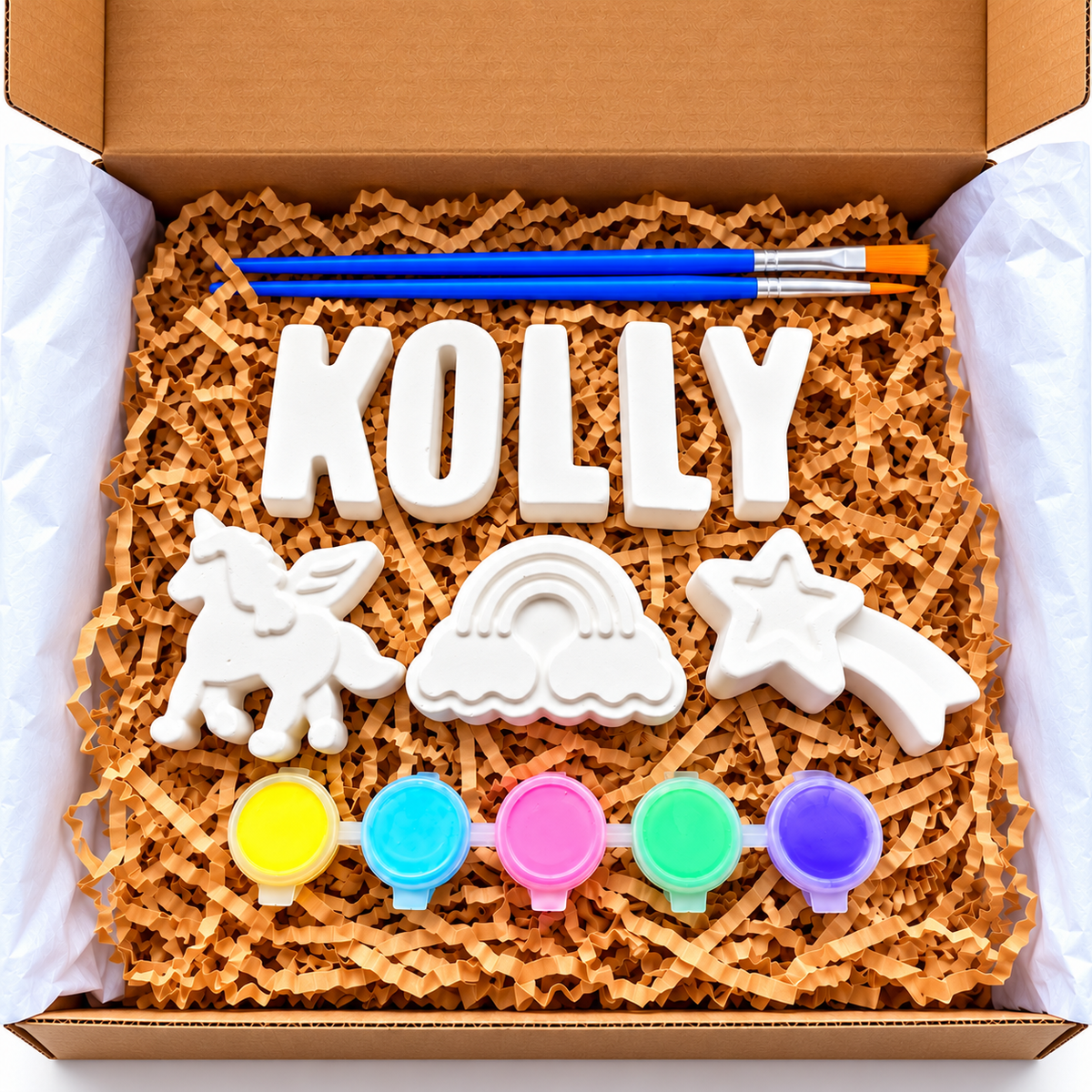 Open cardboard gift box filled with brown crinkle paper, containing a white custom name 'KOLLY' block, a unicorn, a rainbow, a star, five small paint pots, and a paintbrush.