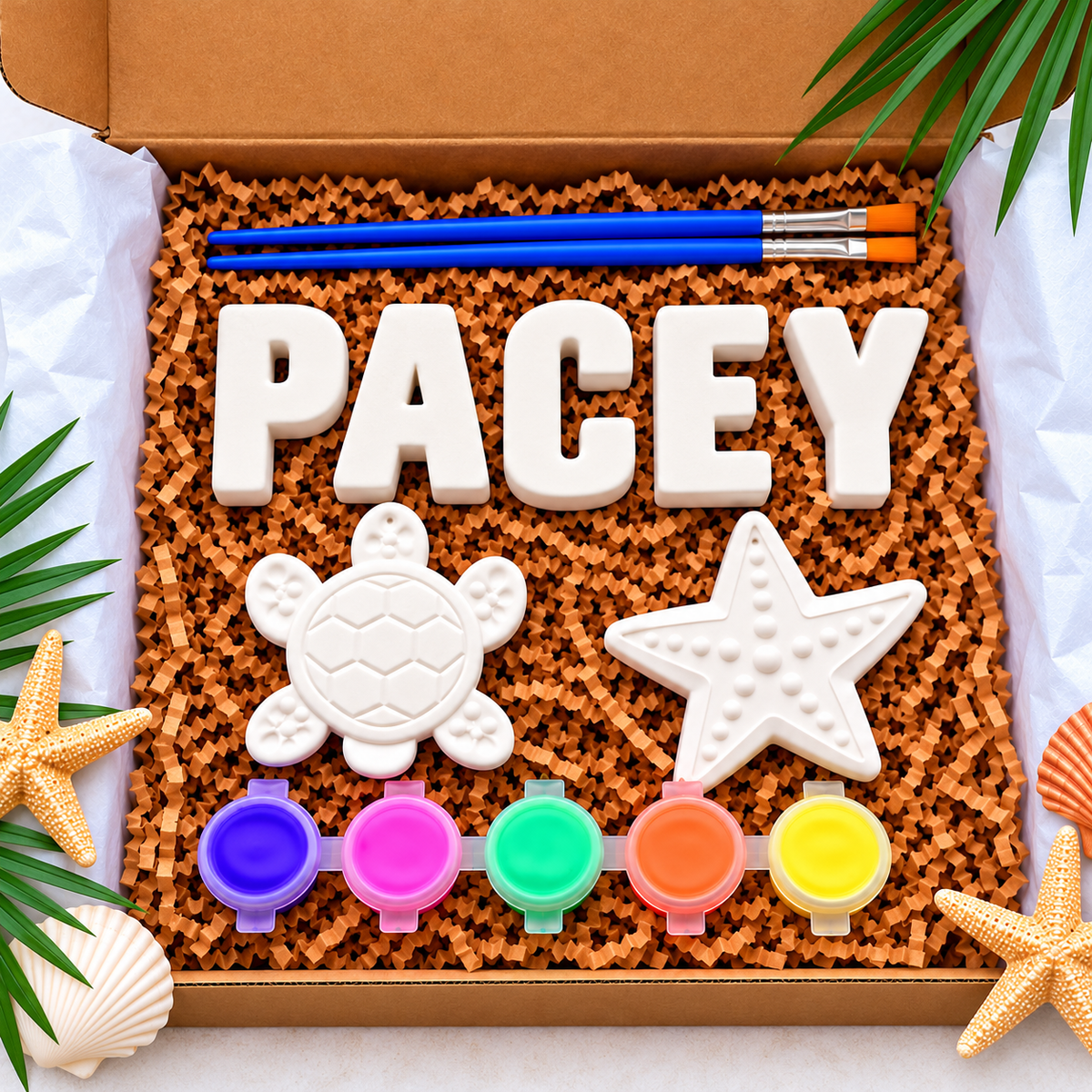 A personalized plaster paint kit in a cardboard box, featuring white plaster letters spelling 'PACEY', a turtle and starfish plaster, five small paint pots, and a blue paintbrush, surrounded by beach-themed props like starfish and seashells.