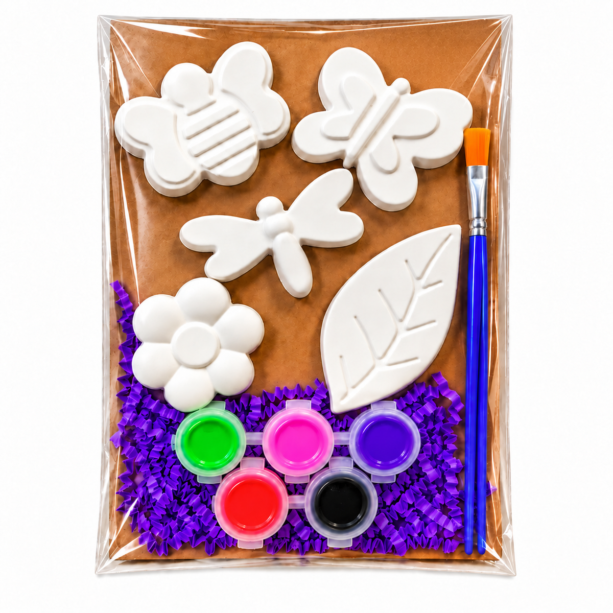A children's painting plaster kit in a clear plastic bag, featuring white plaster shapes of flowers, butterflies, a dragonfly, a leaf, and a bee, along with six small paint pots and a paintbrush on a brown background with purple crinkle paper.